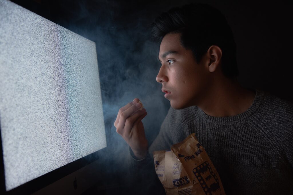 a teenage boy with sad eyes, staring into a blank, fuzzy computer screen while eating popcorn. He appears to be eating alone.  